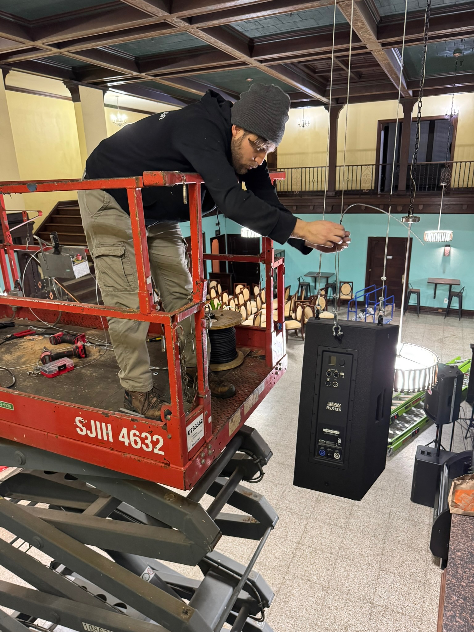 Technician on a lift working on a suspended speaker with the room layout visible below