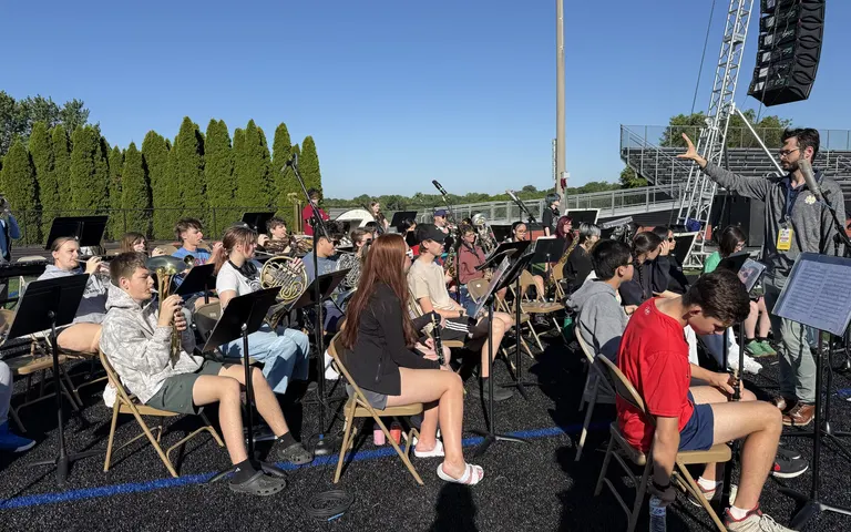 Student band seated on an athletic field beside the outdoor speaker system during graduation preparations