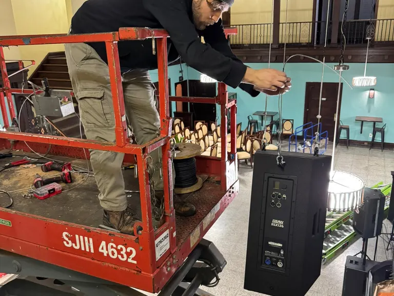 Technician on a lift working on a suspended speaker with the room layout visible below